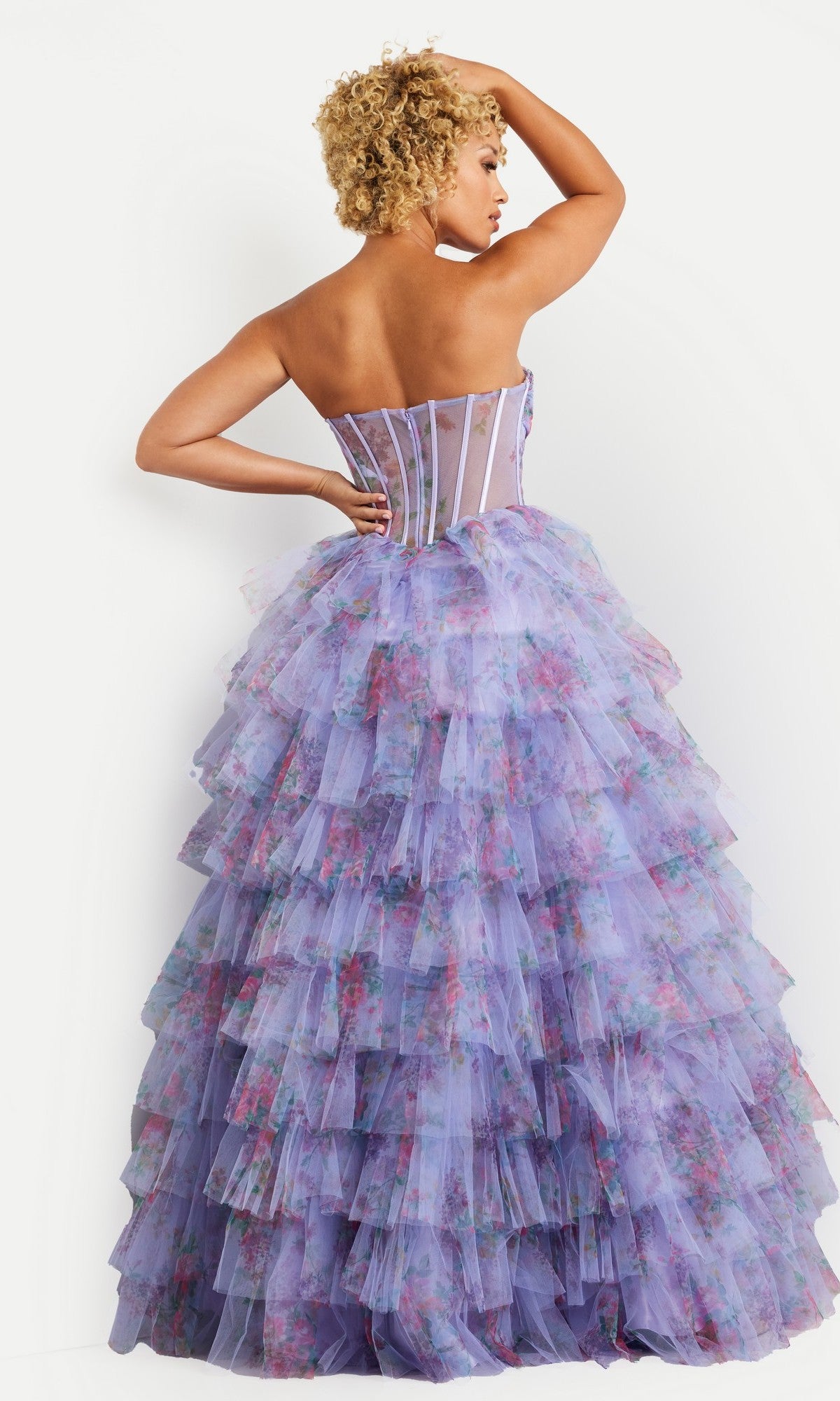 Woman showing the back of a layered, multicolored floral print ball gown dress she is wearing, standing against a white background